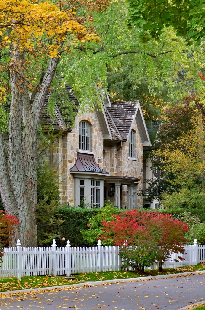 Stone Tudor-style home with white picket fence framed by fall foliage